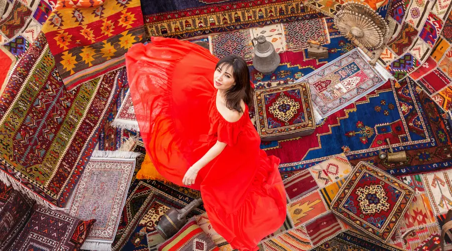 Colorful hand-knotted Kashmiri carpets and rugs with traditional patterns displayed around a woman in a red dress.