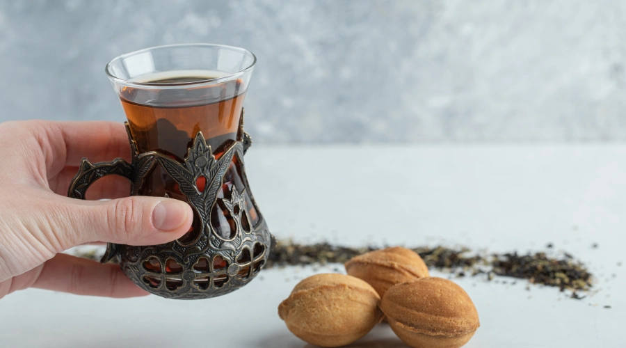 Hand holding a glass cup of Kashmiri Kahwa tea with walnuts and tea leaves on the table.